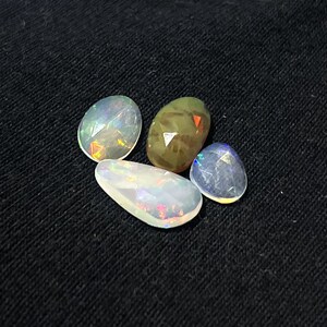 May include: Four faceted gemstones of varying shapes and sizes. Three stones are translucent white with iridescent flashes of color, while one central stone is opaque olive green with subtle orange and red internal light reflections.