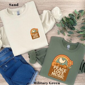 May include: Two sweatshirts, one in sand and one in military green, displaying a retro graphic with a rainbow, dove, guitar, and the words "PEACE LOVE MUSIC." The design is inspired by Woodstock 1969. Blue jeans are also visible.