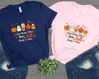 May include: A navy blue t-shirt and a light pink t-shirt, each featuring a graphic of a smiling pumpkin, ghost, coffee cup, maple leaf, and candy corn. The text reads Just Besties Who love Fall, with the names Emily and Anna separated by a heart.