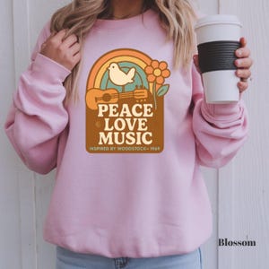 May include: A pale pink sweatshirt with a vintage-style graphic. The design includes a rainbow, dove, guitar, and flower, with the text "PEACE LOVE MUSIC INSPIRED BY WOODSTOCK - 1969". The word "Blossom" is printed in the bottom right corner.