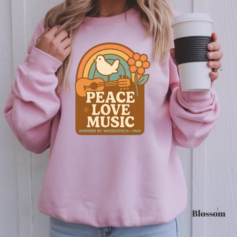 May include: A pale pink sweatshirt with a vintage-style graphic. The design includes a rainbow, dove, guitar, and flower, with the text "PEACE LOVE MUSIC INSPIRED BY WOODSTOCK - 1969". The word "Blossom" is printed in the bottom right corner.