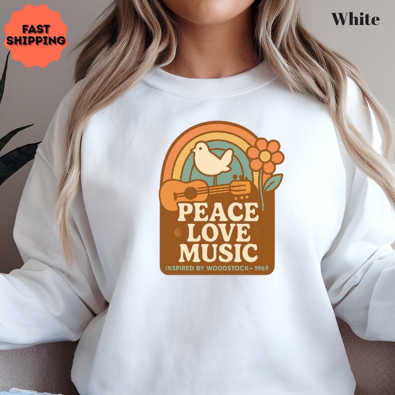 May include: White sweatshirt with a retro design featuring a rainbow, dove, flower, and ukulele. The graphic reads "PEACE LOVE MUSIC" with "INSPIRED BY WOODSTOCK - 1969" below. The sweatshirt has a crew neck.