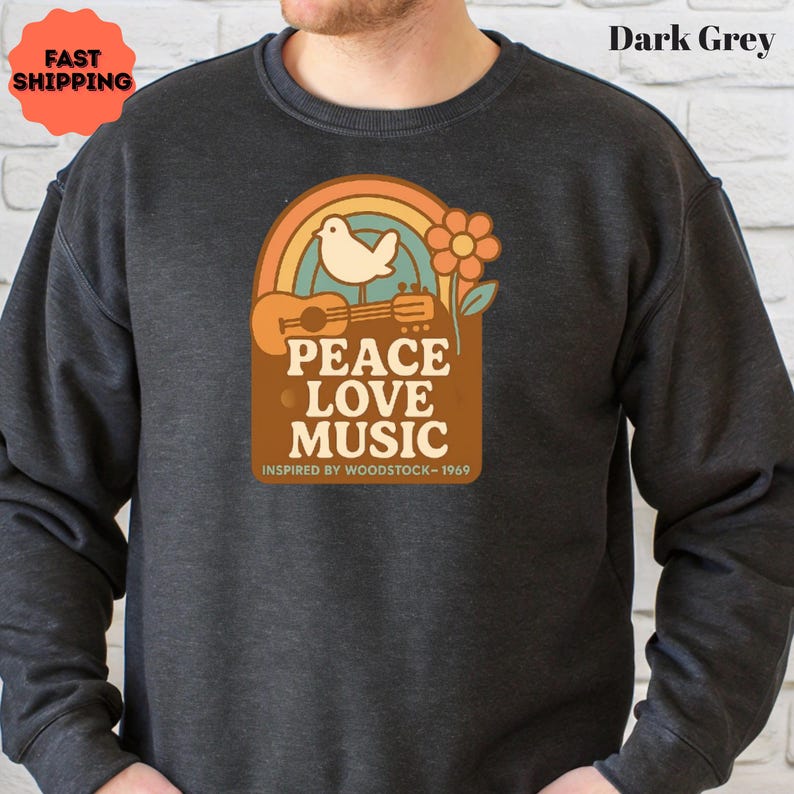 May include: Dark grey sweatshirt with a retro graphic featuring a rainbow, dove, guitar, and flower. The text reads "PEACE LOVE MUSIC" with "INSPIRED BY WOODSTOCK - 1969" below. The sweatshirt has a crew neck and long sleeves.
