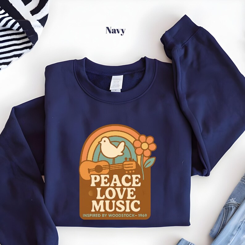 May include: Navy blue sweatshirt with a retro graphic featuring a rainbow, dove, guitar, and flower. The text reads "PEACE LOVE MUSIC" and "INSPIRED BY WOODSTOCK - 1969". A casual, comfortable top for music lovers.