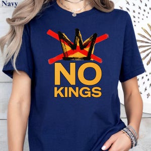 May include: Navy blue t-shirt featuring a graphic design. The design includes a crown crossed out with a red line, and the words "NO KINGS" in bold, yellow letters. The shirt is a short-sleeved crew neck.
