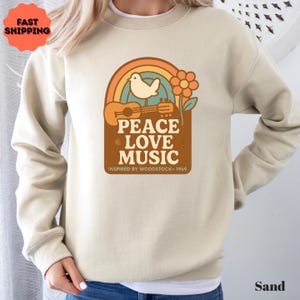 May include: Sand-coloured sweatshirt with a retro graphic featuring a rainbow, dove, guitar, and flower. The text reads "PEACE LOVE MUSIC" and "INSPIRED BY WOODSTOCK - 1969". A vintage-inspired design.