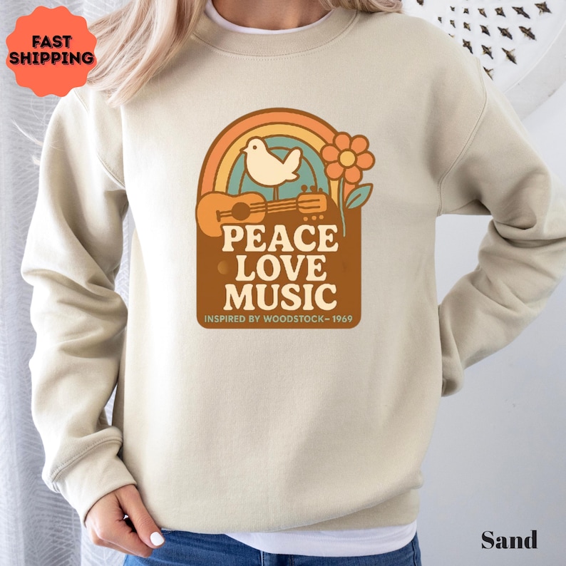 May include: Sand-coloured sweatshirt with a retro graphic featuring a rainbow, dove, guitar, and flower. The text reads "PEACE LOVE MUSIC" and "INSPIRED BY WOODSTOCK - 1969". A vintage-inspired design.