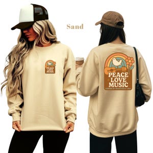 May include: Sand-coloured sweatshirt with a retro graphic on the front and back. The front graphic says "Peace Love Music" with a rainbow and flower design. The back graphic features a rainbow, dove, guitar, and the text "Peace Love Music, Inspired by Woodstock - 1969."