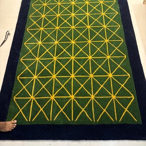 May include: A rectangular rug with a geometric pattern. The rug features a green center with a yellow geometric design and a navy blue border. The geometric pattern consists of interconnected lines forming triangles and squares. The rug is likely made of a soft material.