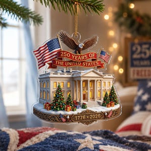 May include: A clear acrylic Christmas tree decoration depicting the White House, American flags, and an eagle. The ornament celebrates "250 Years of The United States" with the years 1776-2026. Christmas trees and gifts are also included in the design.