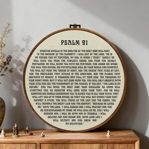 May include: A round, wooden framed wall hanging featuring the text "PSALM 91" and the full text of Psalm 91. The text is in a dark font on a cream-colored background. The frame is a medium brown color.