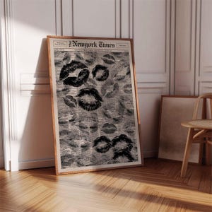 Puede incluir: Un estampado enmarcado con un dise&ntilde;o de peri&oacute;dico vintage con numerosas marcas de besos de l&aacute;piz labial negro. El peri&oacute;dico se titula "New York Times" y se muestra en un marco de madera. El estampado est&aacute; apoyado contra una pared blanca y un suelo de madera.
