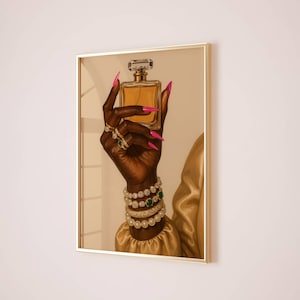 May include: A framed print showing a hand holding a perfume bottle. The hand is dark-skinned, with pink nails, rings, and bracelets. The perfume bottle is gold-toned with a clear stopper. The background is a neutral colour, and the frame is gold.