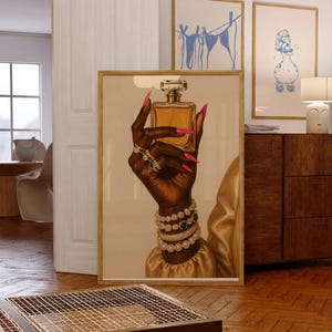 May include: A framed art print showing a dark hand holding a perfume bottle. The hand is decorated with rings, bracelets, and long pink nails. The perfume bottle is square with a clear top. The background is a neutral colour.
