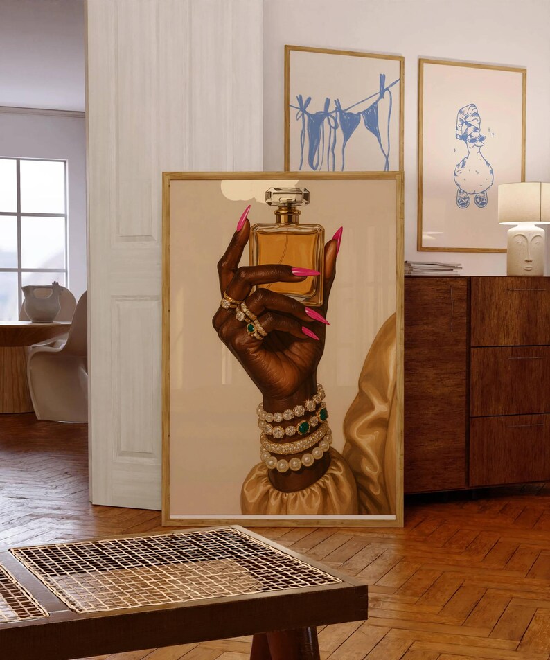 May include: A framed art print showing a dark hand holding a perfume bottle. The hand is decorated with rings, bracelets, and long pink nails. The perfume bottle is square with a clear top. The background is a neutral colour.