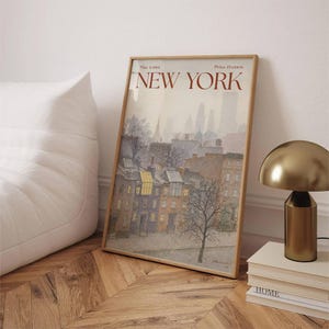 May include: A framed print depicting a New York City street scene in muted colours. The artwork shows buildings, trees, and the text "NEW YORK" in a reddish-brown font. A gold-coloured lamp and a stack of books are next to the print.