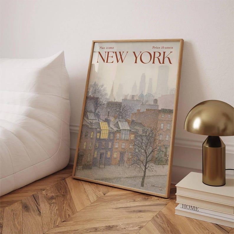 May include: A framed print depicting a New York City street scene in muted colours. The artwork shows buildings, trees, and the text "NEW YORK" in a reddish-brown font. A gold-coloured lamp and a stack of books are next to the print.