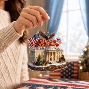 May include: A clear acrylic Christmas ornament depicting the White House, American flags, and an eagle. The ornament celebrates "250 Years of the United States" with the years "1776-2026" displayed. The ornament is held by a person.