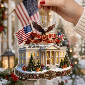 May include: A clear acrylic Christmas ornament depicting the White House, American flags, and an eagle. The ornament celebrates "250 YEARS OF THE UNITED STATES" with the years "1776 * 2026". Decorated with Christmas trees and presents.