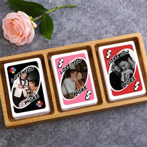 Puede incluir: Una bandeja de madera contiene tres cartas personalizadas con fotos románticas. Las cartas muestran las frases "Love You" y "More" con un símbolo de inversión. Una rosa rosa descansa cerca. Los dorsos de las cartas son negros, rosas y rojos.