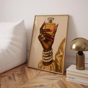 May include: A framed art print depicting a dark hand holding a perfume bottle. The hand is decorated with rings, bracelets, and long pink nails. The artwork has a beige background and a gold frame. The image conveys a sense of luxury and beauty.