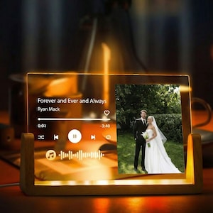 Może przedstawiać: Przezroczysta akrylowa ramka na zdjęcia z drewnianą podstawą, wyświetlająca zdjęcie ślubne i interfejs odtwarzacza muzyki. Tekst brzmi "Forever and Ever and Always" autorstwa Ryana Macka. Ramka jest podświetlona ciepłym, złotym światłem.