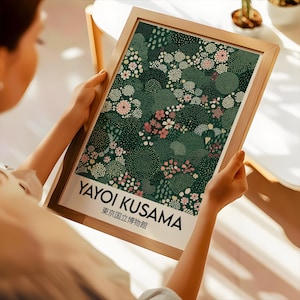 May include: A framed art print featuring a floral design in shades of green, pink, and white. The print includes the text "YAYOI KUSAMA" and Japanese text. The frame is made of light-colored wood. The artwork is held by a person.
