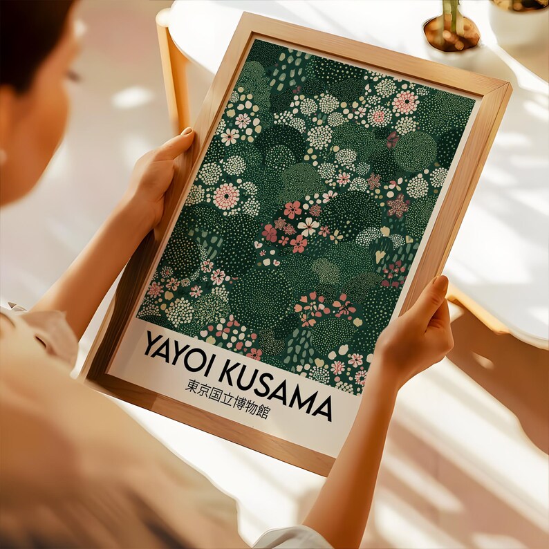 May include: A framed art print featuring a floral design in shades of green, pink, and white. The print includes the text "YAYOI KUSAMA" and Japanese text. The frame is made of light-colored wood. The artwork is held by a person.
