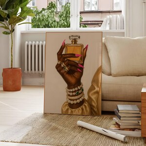 May include: An art print showing a dark hand holding a gold perfume bottle. The hand is decorated with rings, bracelets, and long pink nails. The artwork is set against a neutral background, with a gold sleeve visible.