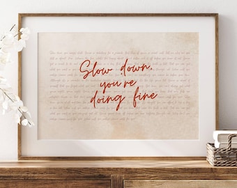 May include: A framed print with the words "Slow down, you're doing fine" in red script. The background is a beige color with a handwritten style font. The frame is a light brown color.