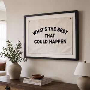 May include: A rectangular wall art piece featuring a cream-colored fabric banner with black text that reads WHAT'S THE BEST THAT COULD HAPPEN. The banner is secured with two brass-colored metal grommets and is encased in a black wooden frame.