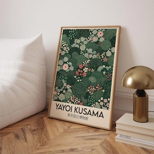 May include: A framed art print featuring a floral pattern in shades of green, pink, and white. The print includes the text "YAYOI KUSAMA" and Japanese text. The artwork is displayed on a wooden floor, next to a gold lamp and books.