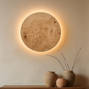 May include: A modern wooden console table with a round, illuminated wall art piece. The table features a wooden top supported by large, rounded wooden bases. Three ceramic vases and branches sit on the table.