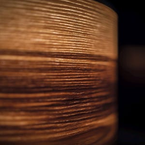 May include: A cylindrical object with a visible wood grain texture featuring horizontal lines in shades of light and dark brown. The surface displays a natural pattern of wood fibers with a smooth finish, captured in a side profile view.