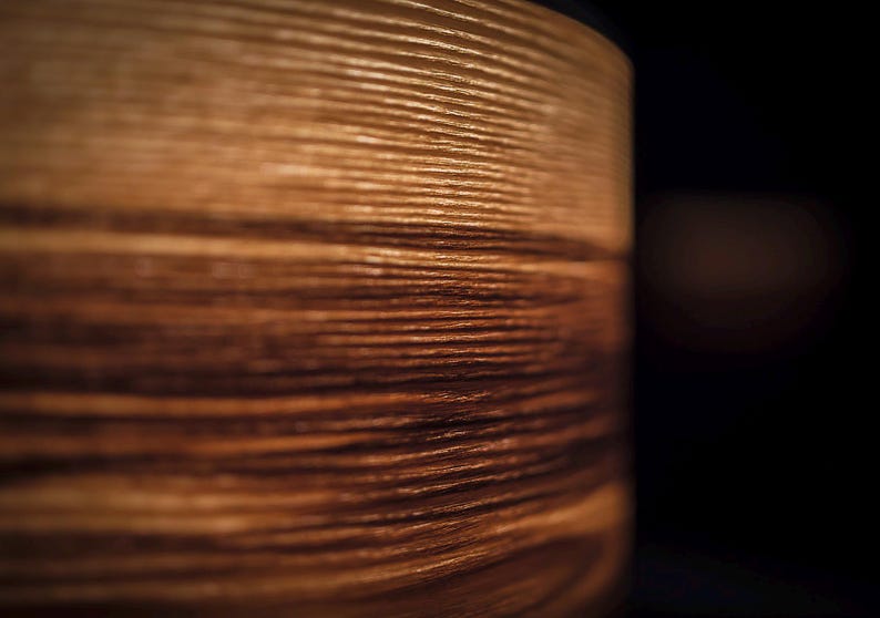 May include: A cylindrical object with a visible wood grain texture featuring horizontal lines in shades of light and dark brown. The surface displays a natural pattern of wood fibers with a smooth finish, captured in a side profile view.