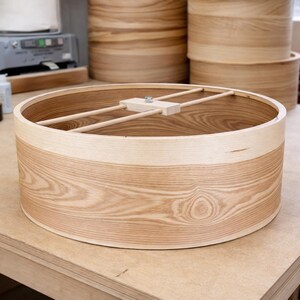 May include: A circular light-colored wooden drum shell featuring a prominent natural grain pattern. The interior includes a central wooden support bar with a metal bolt mechanism spanning the diameter of the open cylinder structure.