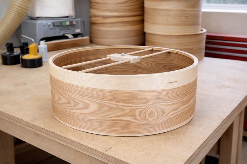 May include: A circular light-colored wooden drum shell featuring a prominent natural grain pattern. The interior includes a central wooden support bar with a metal bolt mechanism spanning the diameter of the open cylinder structure.