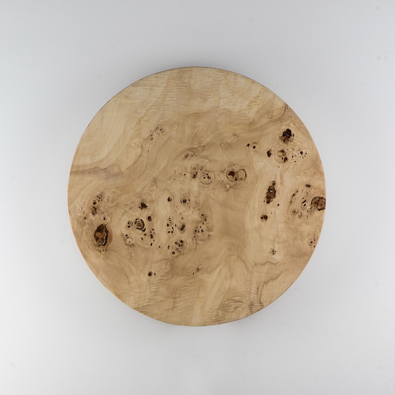 May include: A round wooden serving board with a light brown color and a unique wood grain pattern. The board has a natural, organic look, with small knots and variations in the wood. The board is perfect for serving appetizers or as a decorative piece.