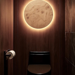 May include: A modern bathroom interior featuring a black toilet and a round wooden wall light with a warm, glowing perimeter. The walls are paneled with dark wood, creating a luxurious and inviting atmosphere. A roll of toilet paper is mounted on the wall.