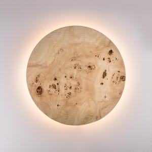 May include: A round, wooden wall sconce with a natural wood grain pattern. The sconce has a warm, neutral color palette with dark brown knots and a soft, glowing light emanating from behind the wooden disc. The design is modern and minimalist.