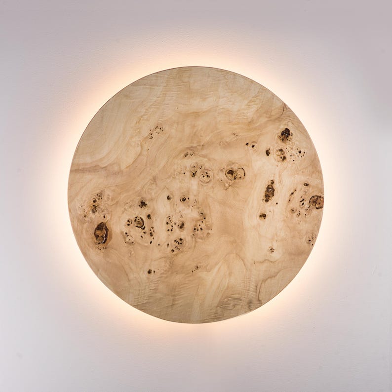 May include: A round, wooden wall sconce with a natural wood grain pattern. The sconce has a warm, neutral color palette with dark brown knots and a soft, glowing light emanating from behind the wooden disc. The design is modern and minimalist.