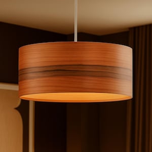 May include: A cylindrical pendant light fixture with a warm wood grain finish. The lampshade has a dark band of wood grain around the middle. The light fixture hangs from a white cord and illuminates the room with a soft glow.