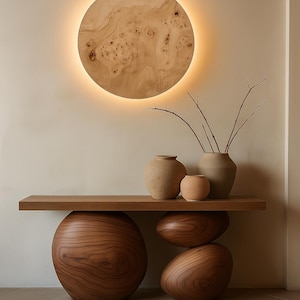May include: A modern wooden console table with a round, illuminated wall art piece. The table features a wooden top supported by large, rounded wooden bases. Three ceramic vases and branches sit on the table.