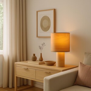 May include: A wooden table lamp with a cylindrical base and a lampshade. The lamp sits on a light wood table with two drawers. A framed print with a circular design hangs on the wall. Decorative vases and a bowl are on the table.