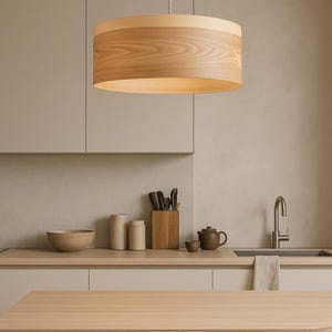 May include: A cylindrical pendant light fixture featuring a light-toned wood veneer shade with a visible natural grain pattern. The drum-shaped shade is suspended from a white cord, providing downward illumination in a neutral-colored interior space.