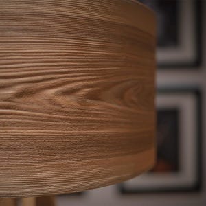 May include: A cylindrical lamp shade featuring a light brown wood grain texture. The surface displays horizontal lines and organic patterns that mimic the appearance of natural timber, creating a tactile and detailed visual effect on the exterior.