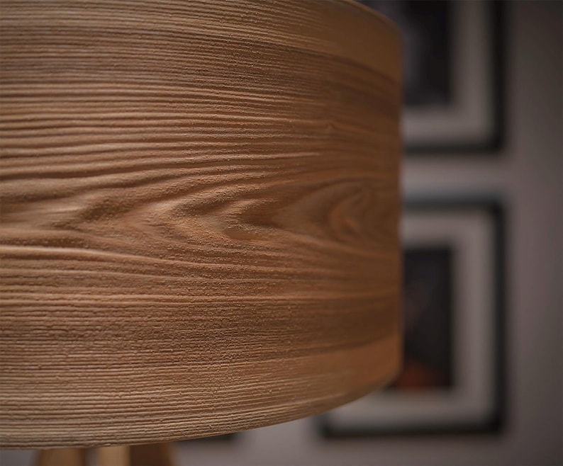 May include: A cylindrical lamp shade featuring a light brown wood grain texture. The surface displays horizontal lines and organic patterns that mimic the appearance of natural timber, creating a tactile and detailed visual effect on the exterior.