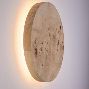 May include: A wooden wall sconce with a circular design. The sconce is made of light-colored wood with a natural grain pattern. A warm light emanates from behind the wooden disc, creating a soft glow. The sconce is mounted on a white wall.