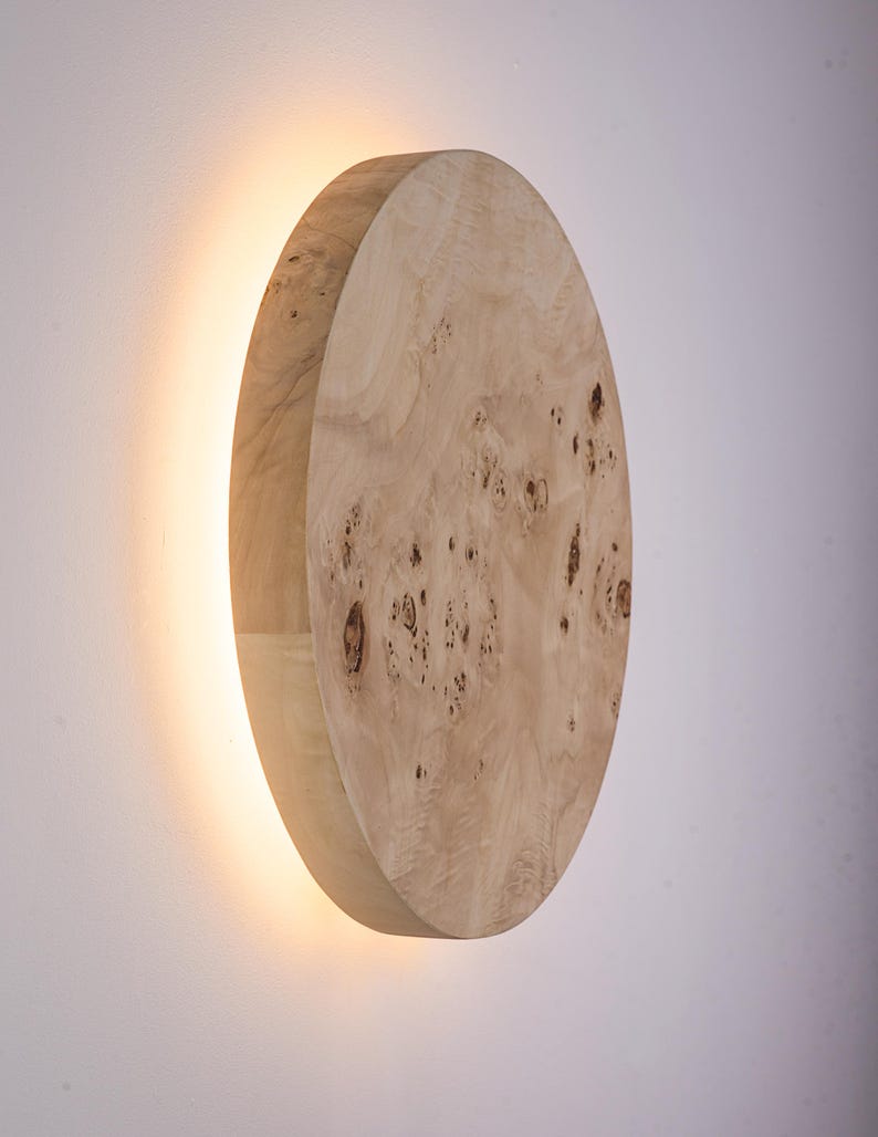 May include: A wooden wall sconce with a circular design. The sconce is made of light-colored wood with a natural grain pattern. A warm light emanates from behind the wooden disc, creating a soft glow. The sconce is mounted on a white wall.