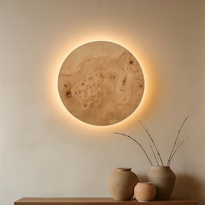 May include: A round, wooden wall light fixture with a warm, glowing halo. The wood grain is visible, and the fixture is mounted on a neutral-colored wall. Decorative vases and branches are in the foreground.